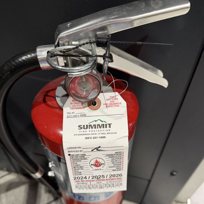 Overhead view of extinguisher with a Summit Fire Protection inspection tag containing fields for license number, who it was serviced by, and punch holes for type of inspection and inspection date