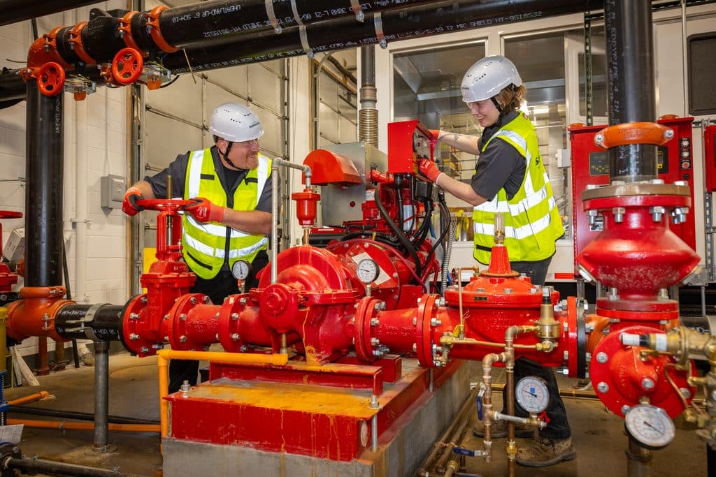 Two Summit technicians wearing hard hats and high-visibility vests work on a red fire pump assembly and its controls in a mechanical room.