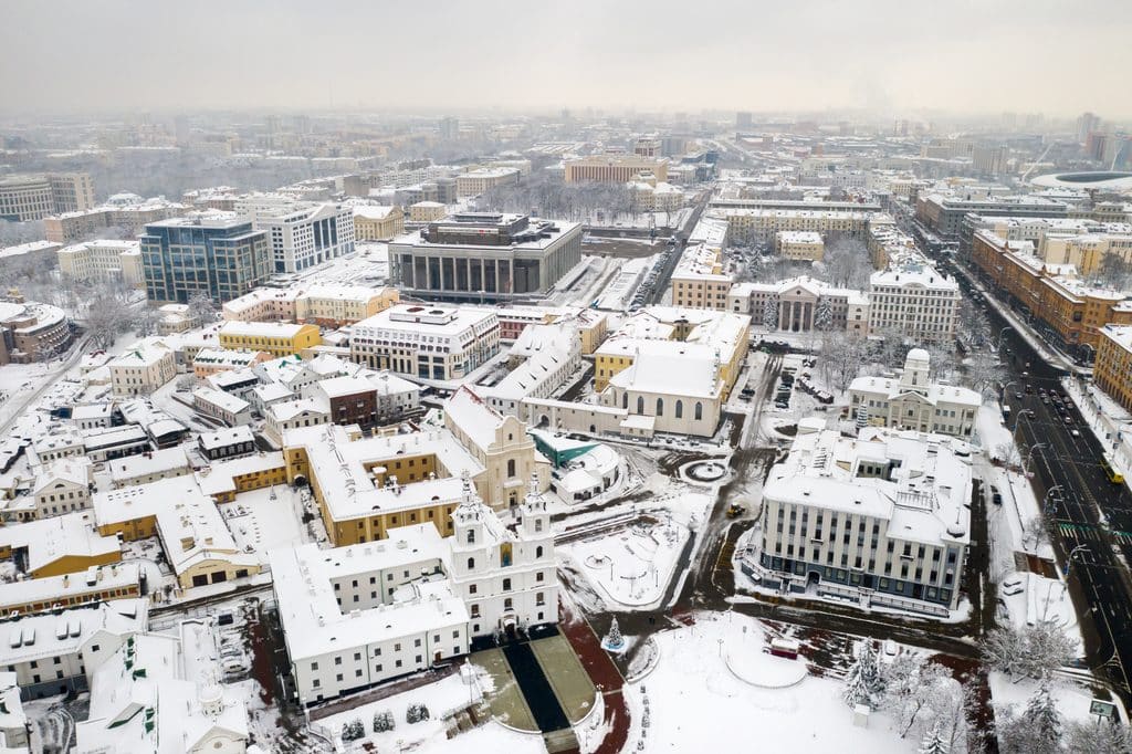 An eye-level view captures a city covered in snow during winter. Buildings of varying sizes and architectural styles are seen. The sky is overcast, creating a soft, diffused light across the wintry scene. The scene evokes a sense of cold and peace, and could be used for projects centered on architecture, travel, winter, or cityscapes.